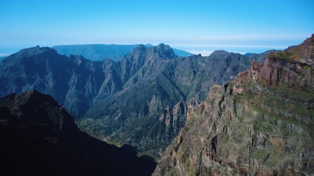 Aerial panning shot of majestic volcanic cliffs at Pico do Areeiro, drone rotating right to left revealing red basalt ridges and green highlands above the clouds in Madeira, cinematic.