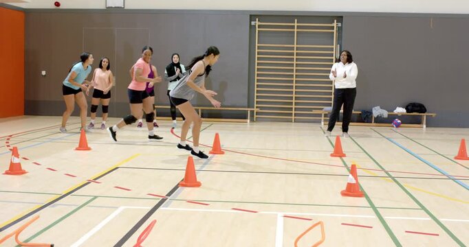 With coach clapping, diverse female athletes stepping through rope jumping cones sprinting on court