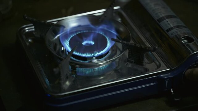 Close-up shot of a portable blue gas stove igniting with a burst of orange fire settling into a steady circular blue flame on a dark background with sound