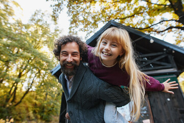 Fototapeta premium A father gives his daughter a piggyback ride in a colorful autumn forest. Their laughter fills the air as golden leaves surround them, creating a perfect weekend memory.