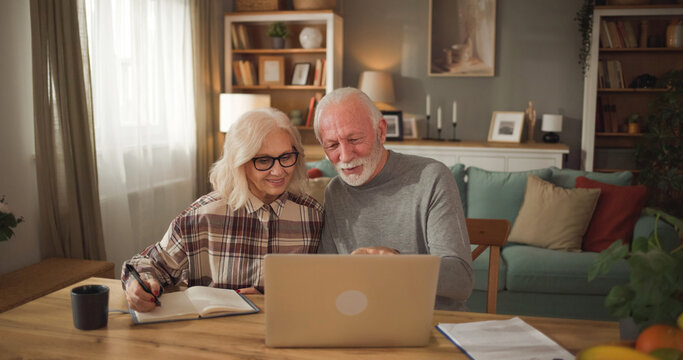An elderly couple sits at a wooden table, looking at a laptop together. They are discussing life insurance options and taking notes in a notebook, surrounded by a cozy home setting.
