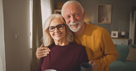 An older couple stands in a comfortable living room holding mugs. They smile joyfully at each other while posing for a picture. Natural light comes through the window. © Stockphotodirectors