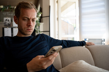 Blonde man sits on a comfortable sofa, deeply focused on his mobile device screen. His furrowed brow shows a thoughtful or concerned mood while relaxing indoors during the day. © Mads