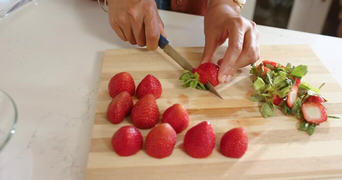 Hands are entering frame, slicing strawberries with blue knife on wooden board, preparing to serve