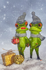 Two green frogs walking hand in hand in the snow and wearing scarves and hats