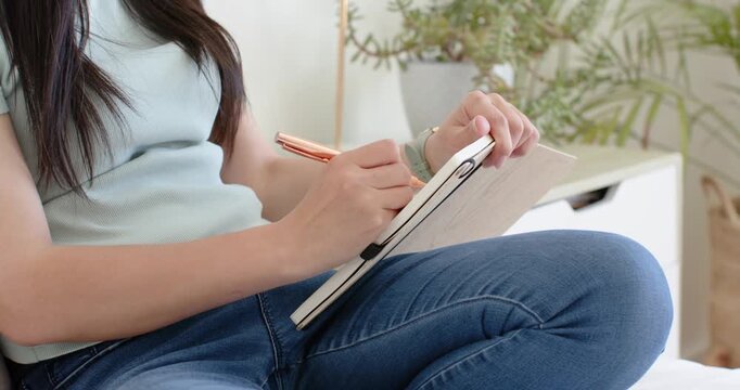 After placing hardback journal on right thigh on bed, she is writing with rose-gold pen, planning
