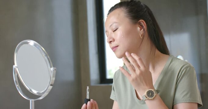 Asian woman lifting dropper, applying serum, massaging face with roller at vanity for skincare