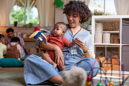 Caregiver holds baby, plays with toy in daycare setting.