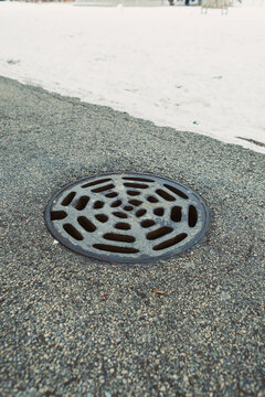 Close view of a circular metal drain cover on a paved road near snow on a winter day in an urban area with no people in sight