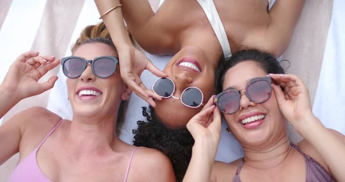 Three women in swimwear lying on striped towel, center lowering sunglasses, adjusting and relaxing