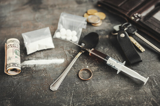 Concept of drug addiction, broken life and ruined marriage, still life with syringe, white powder, pills, empty wallet, coins, rolled banknotes, keys and wedding ring on dark surface