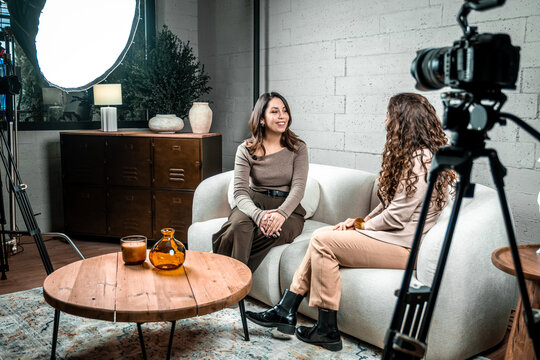 Two women discussing during a video production, sitting on a couch in a modern studio with a camera and lighting setup