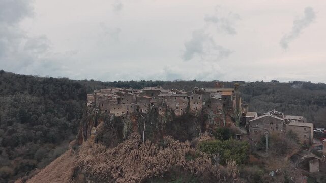 Aerial view of Calcata Vecchia, an ancient medieval village perched on a rocky cliff in Lazio, during a gloomy day. Historic stone houses surrounded by forest and dramatic landscape of a scenic view.
