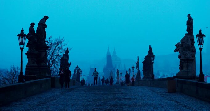 Quiet winter morning on Charles Bridge in Prague with only a few people crossing. Cold blue atmosphere and soft dawn light create a peaceful and cinematic city scene.