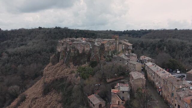 Aerial view of Calcata Vecchia, an ancient medieval village perched on a rocky cliff in Lazio, during a gloomy day. Historic stone houses surrounded by forest and dramatic landscape of a scenic view.
