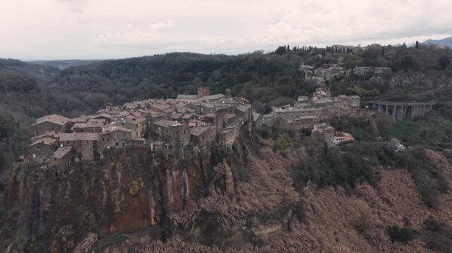 Aerial view of Calcata Vecchia, an ancient medieval village perched on a rocky cliff in Lazio, during a gloomy day. Historic stone houses surrounded by forest and dramatic landscape of a scenic view.
