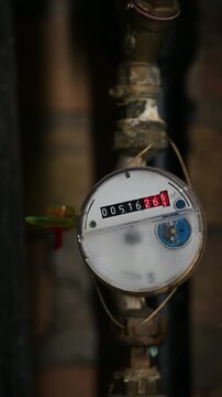 Close-up view of a residential analog water meter connected to vertical rusted pipes in a dimly lit utility area showing consumption dials and numbers