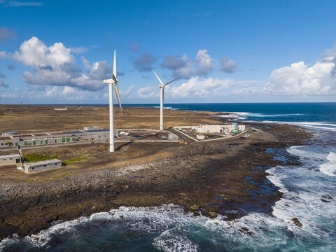 Aerial view of wind turbines standing tall against the crashing waves, powering the grid on the rocky coast, Corralejo, Fuerteventura, Spain.