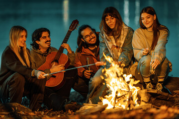 Fototapeta premium Friends gather around a campfire at night by the water enjoying music and roasting marshmallows in the great outdoors