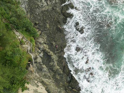Aerial view of the rugged coastline where emerald waters crash against the dark with angler, rocky shore near the lush greenery and stairs of Siung Beach, Gunungkidul Regency, Indonesia.