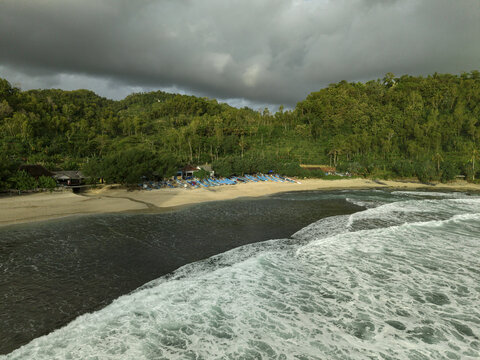 Aerial view of white-crested waves meeting the sandy shore, backed by lush green hills under a dramatic sky, Siung Beach, Gunungkidul Regency, Special Region of Yogyakarta, Indonesia.