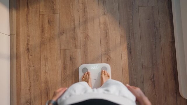 Top view of a young woman stepping onto a smart scale in a bright modern home. Morning routine moment focused on health, body tracking, and mindful weight management. Wooden floor and soft daylight