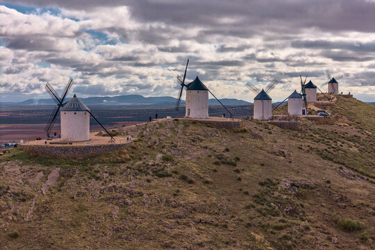 Aerial view of stark white windmills punctuate the rugged, ochre-toned landscape under a dramatic, cloudy sky, iconic against the distant horizon, Consuegra, Castile-La Mancha, Spain.