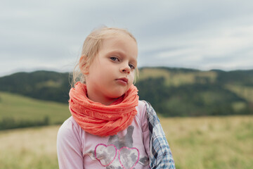Pensive Toddler Girl in Orange Flowy Scarf and Pink Heart Shirt Gazes Thoughtfully over Green Rolling Hills under Cloudy Sky