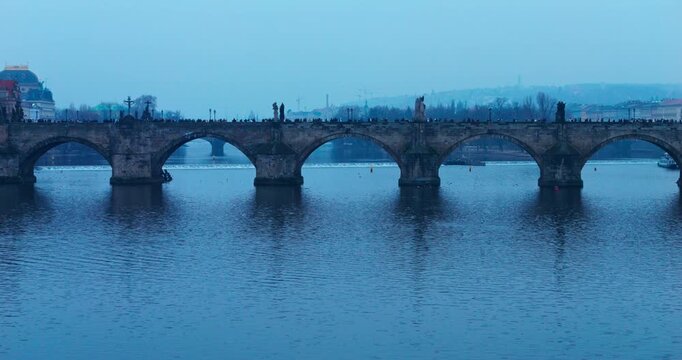 Cold blue winter morning in Prague with a distant view of Charles Bridge over the Vltava River. Tourists cross the historic landmark in a calm, frosty atmosphere.
