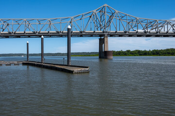 A wide shot shows a large metal truss bridge spanning a wide, calm river under a clear blue sky.