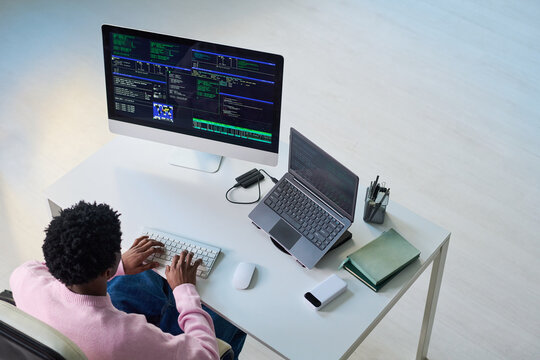 Young Black man working on computer and laptop at desk, typing code on keyboard, multiple monitors displaying programming interfaces, top view workspace