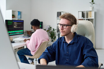Obraz premium Young man wearing headphones working on computer at desk in office, Black young man sitting at adjacent workstation typing on keyboard, both focused on coding tasks