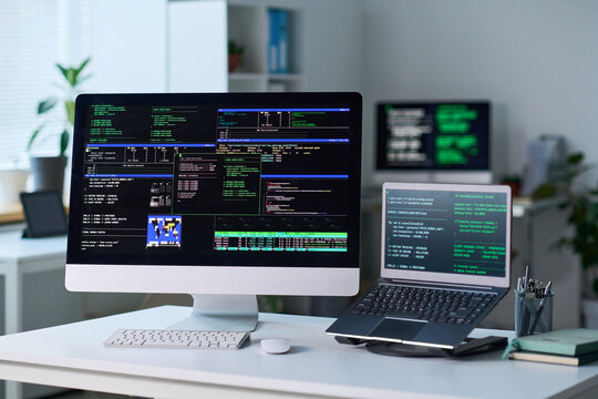 Office workspace showing desktop computer and laptop displaying programming code on screens, digital devices positioned on desk in bright professional environment