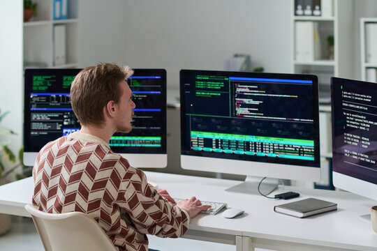 Young man working at desk using multiple computer monitors displaying programming code and system data, focusing on software development in office setting