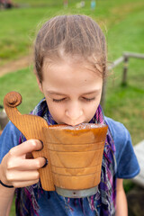A delighted little girl with her eyes closed tries Zentyca - a traditional, thick drink made from sheep's whey, made in Podhale from a wooden ladle and a purple scarf, on a bench in a meadow