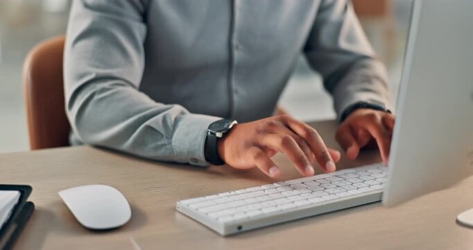 Close-up of person of color typing on white keyboard wearing light gray button-up shirt with wristwatch, seated at wooden desk in evening office with soft warm lighting and blurred background