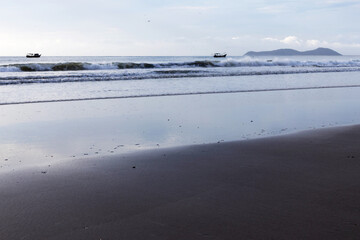 A strip of wet sand forming a mirror-like surface, followed by waves breaking on the beach and two boats at sea - Landscape on the southern coast of S&atilde;o Paulo