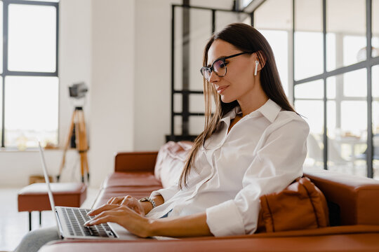 woman sitting on sofa at home relaxed using laptop in white t-shirt and jeans listening to earpods