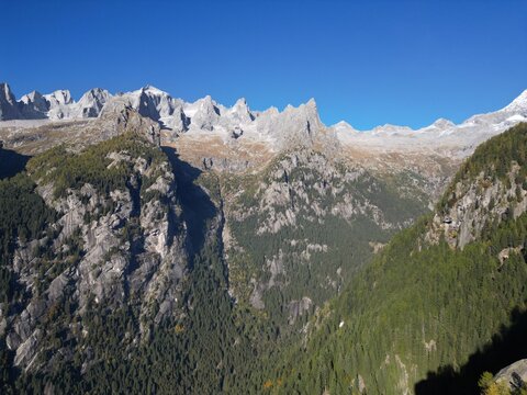 Aerial view of the rugged Val di Mello valley under a clear sky, where rocky cliffs meet dense forests in a dance of light and shadow, Sondrio, Lombardia, Italy.