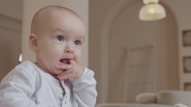 Caucasian baby sitting on bed and putting fingers in mouth. Thoughtful wide eyes and gentle chewing motion. Neutral tones of the nursery and soft authentic lighting. 
