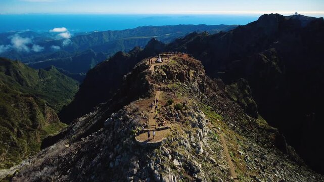 Aerial flyover of Pico Ruivo summit, drone flying over hikers and geodesic marker towards distant Pico do Areeiro observatory, cinematic mountain landscape with ocean horizon in Madeira, 4K.