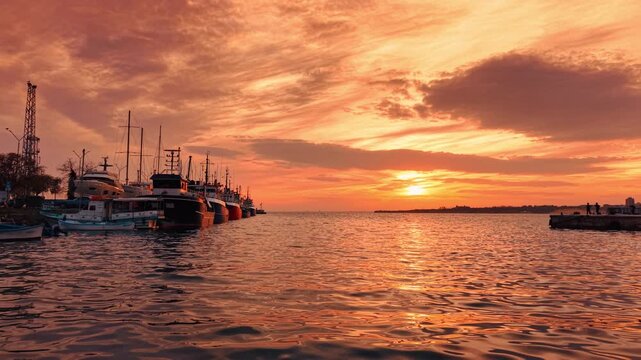 Moored yachts and ships in port at sunset light. Row of boats and ships docked at a stone pier with people walking in the distance under a glowing sunset sky.