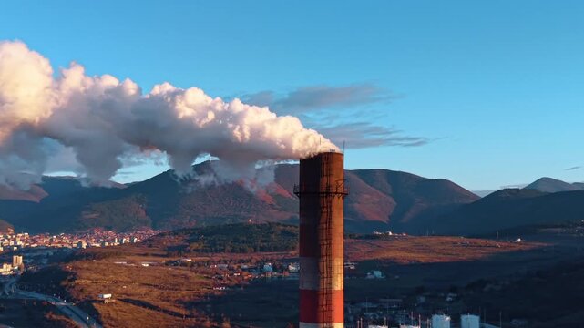Industrial smokestack with thick smoke plume against mountains. Tall red and white chimney emitting dense smoke at sunset with mountain landscape and city in background.