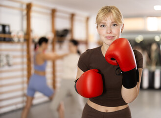 Naklejka premium Girl coach in boxing gloves is standing in gym, students are fighting in background. Active training, sports as lifestyle, active hobbies