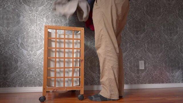 Man wearing khakis putting dirty clothes in a wooden close hamper with a hardwood floor and patterned wall paper