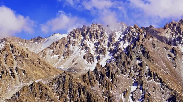 Rocky mountain peaks rise into the sky, with patches of snow visible. Clear blue sky and clouds enhance the wild and serene atmosphere. Drone view.