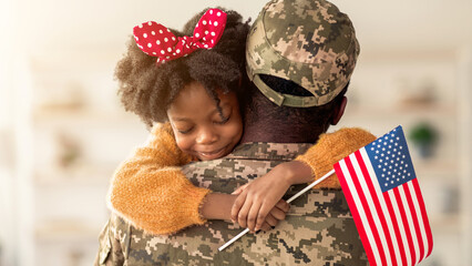 A child with curly hair and a red bow hugs a soldier wearing a camouflage uniform. The soldier holds an American flag. This moment shows joy and warmth during a homecoming event. © Prostock-studio