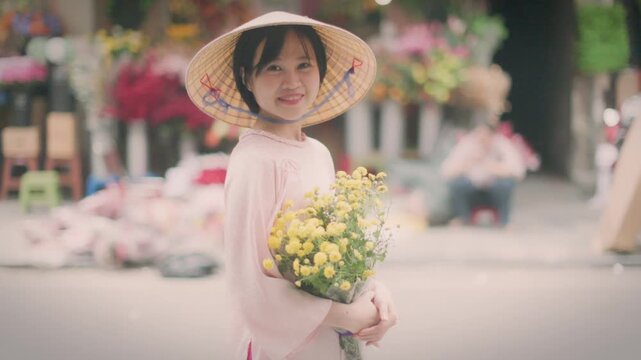 A young woman in a traditional conical hat smiles while holding yellow flowers at a flower market in Old Quarter, Hanoi, Vietnam. She is buying flowers for Tet.