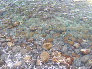 Transparent, crystal-clear waters of the Mediterranean Sea revealing smooth pebbles and stones on the seabed along the coast of Calabria, southern Italy. Sunlight reflects through the shallow water