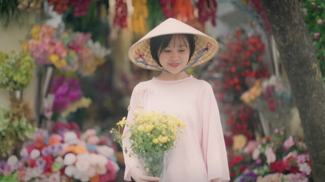 A young woman in a traditional conical hat holds yellow flowers at a flower market in Old Quarter, Hanoi, Vietnam. She is smiling and wearing a pink dress, surrounded by colorful blooms.
